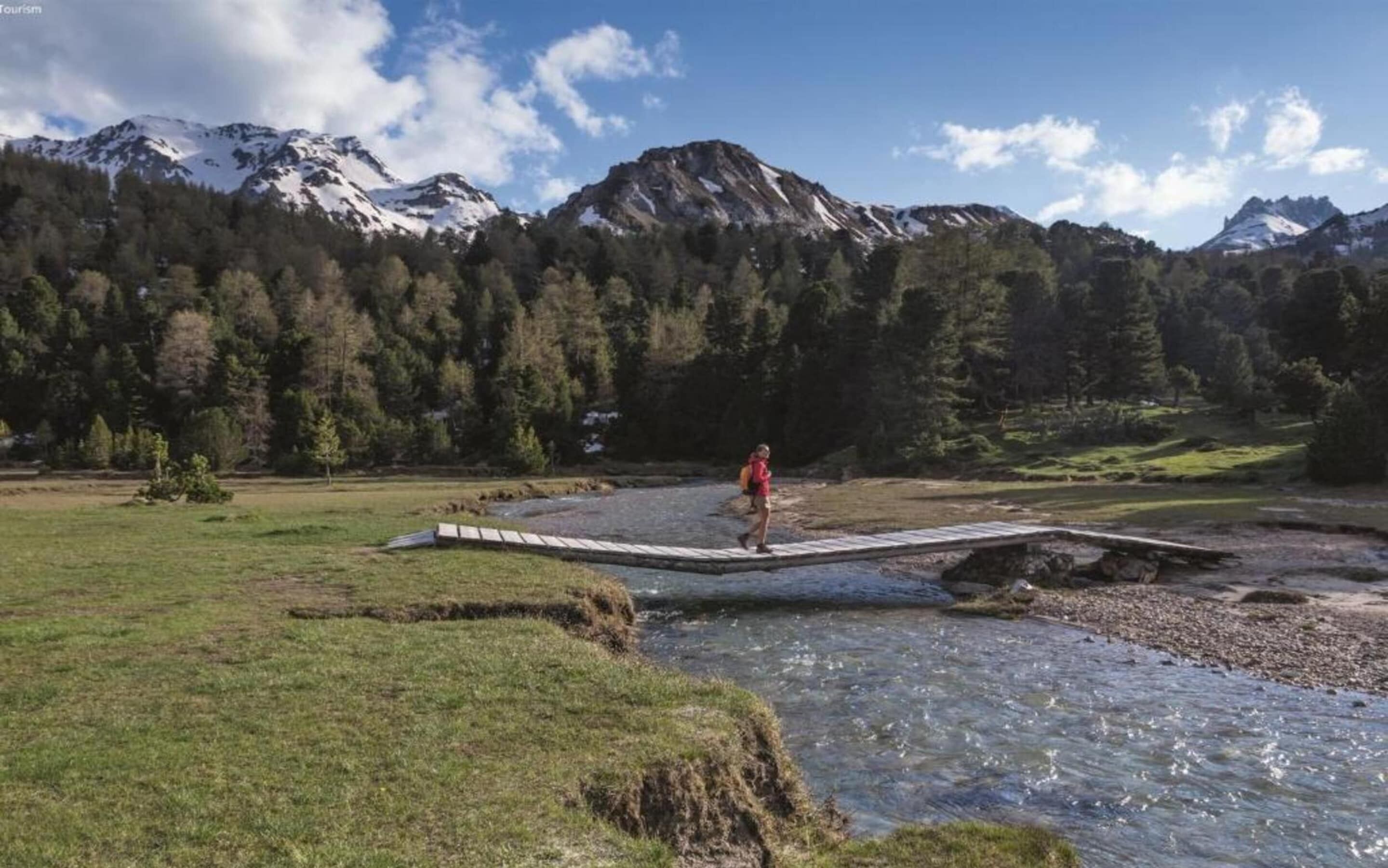 Eine Person überquert eine Brücke über einem Fluss in einer gebirgigen Landschaft, im Hintergrund schneebedeckte Gipfel.