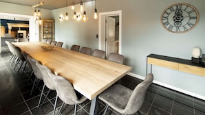 Een moderne eetruimte met een lange houten tafel, grijze gestoffeerde stoelen, hanglampen en een grote wandklok.