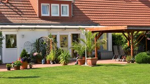 Ein zweistöckiges Haus mit rotem Ziegeldach, einer überdachten Veranda mit Sitzgelegenheiten und einem gepflegten Garten mit verschiedenen Kübelpflanzen.