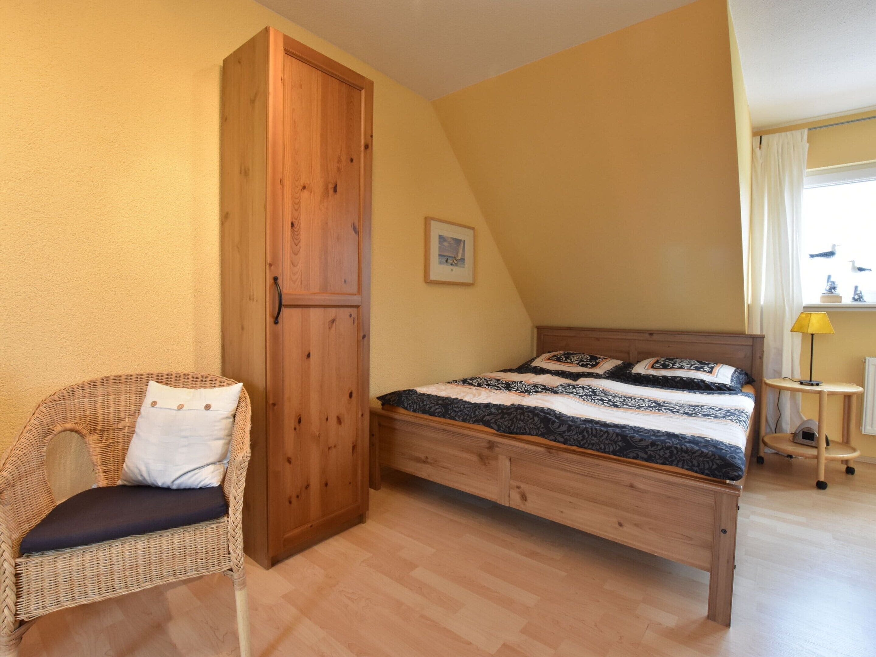 Ein Schlafzimmer mit einem Bett, einem Korbstuhl, einem Holzschrank und einem kleinen Tisch mit einer Lampe.