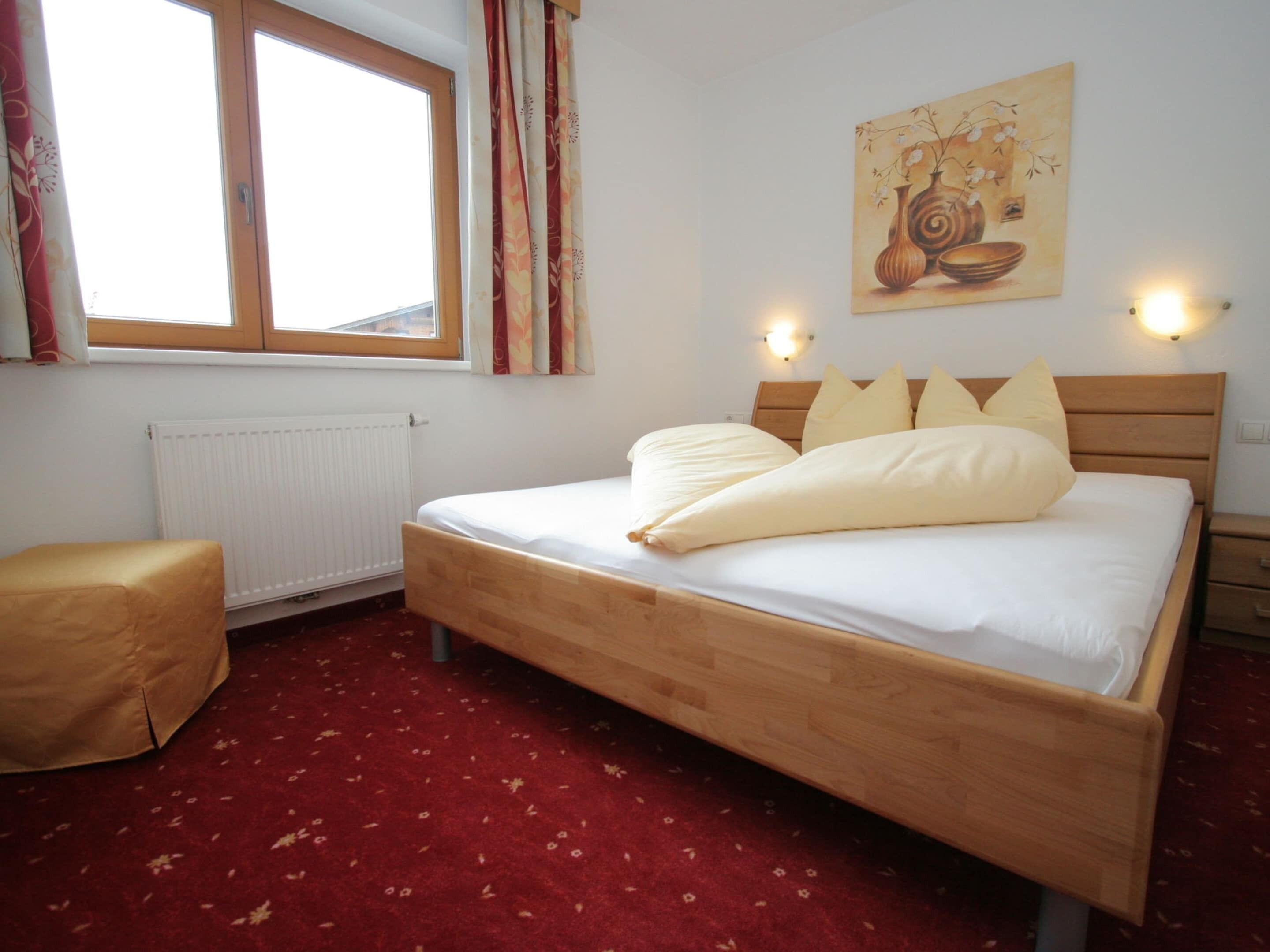 Une chambre avec un lit en bois, une moquette rouge, une fenêtre avec des rideaux, un radiateur et un tableau accroché au mur.