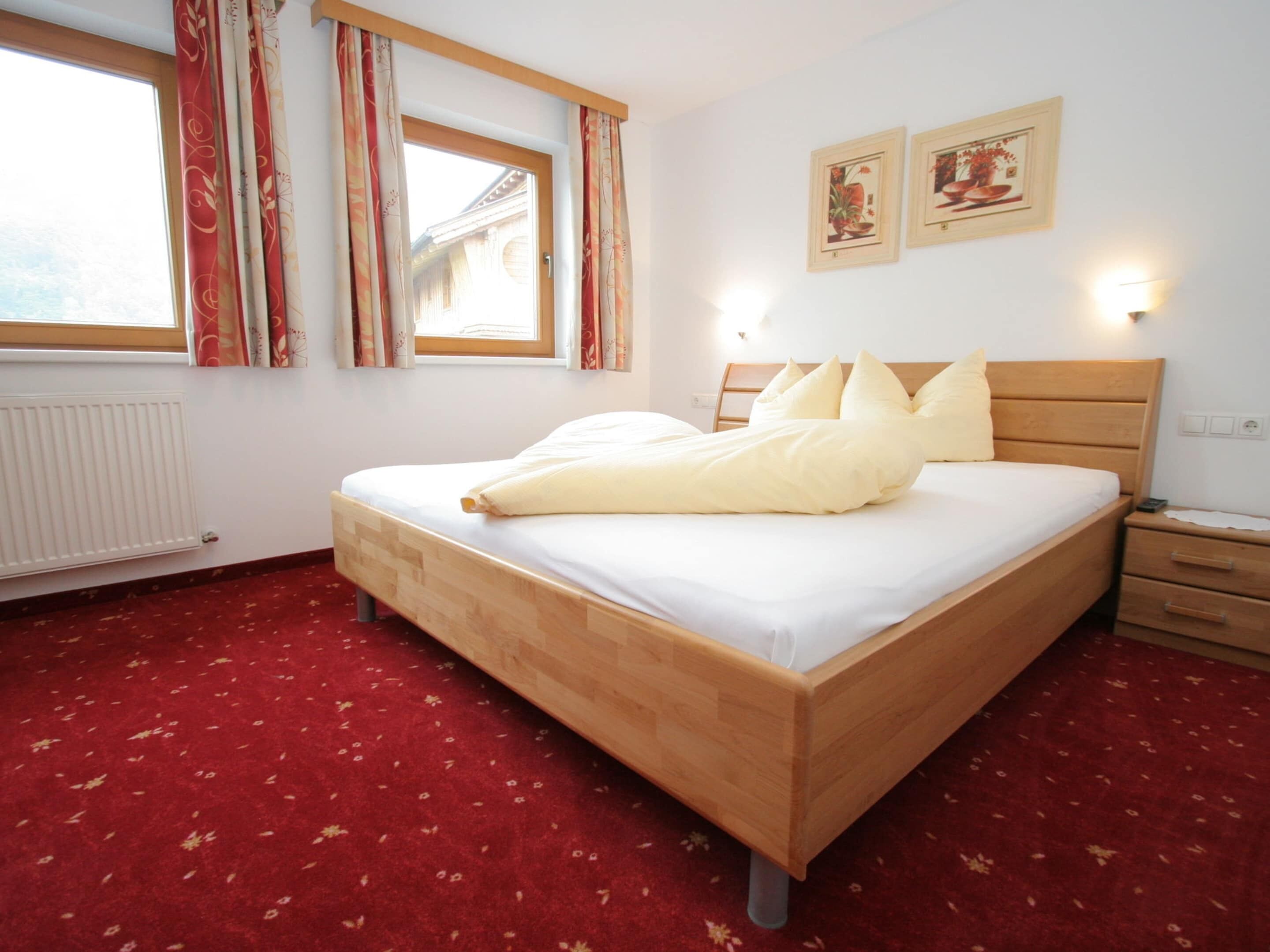 A neatly made bed with a wooden headboard, a red carpeted floor, a window with curtains, and two wall-mounted light fixtures.