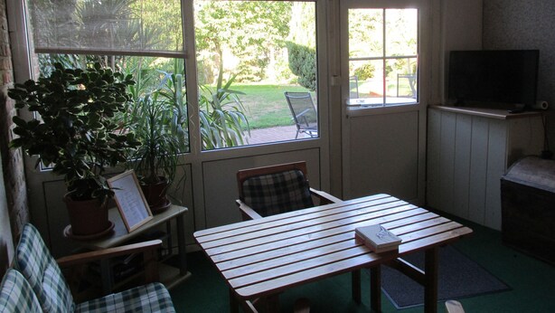 Une pièce chaleureuse avec une table en bois, un fauteuil à carreaux, un téléviseur posé sur un meuble et une grande fenêtre donnant sur un jardin.