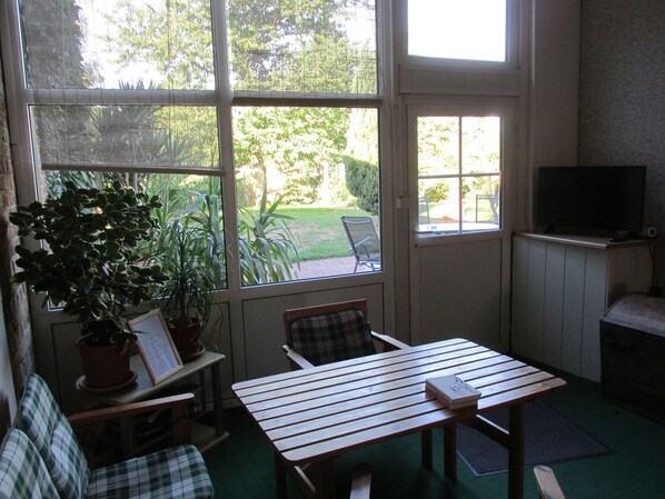 Um quarto aconchegante com uma mesa de madeira, uma cadeira xadrez, uma televisão em um suporte e uma grande janela com vista para um jardim.