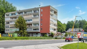 Un edificio de varios pisos con balcones, una esquina roja y una calle con coches y una parada de autobús.
