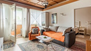 Una sala de estar acogedora con un sofá, una mesita y una silla. Hay una ventana con cortinas, un ventilador de techo y un perchero.