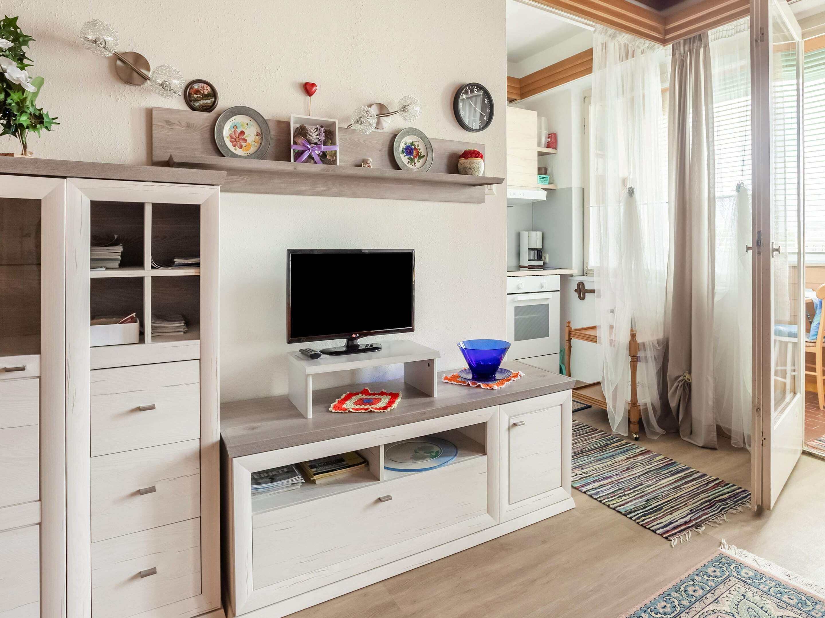 A living room with a TV, shelves with decorative items, a blue bowl, and a kitchen area visible through an open door.