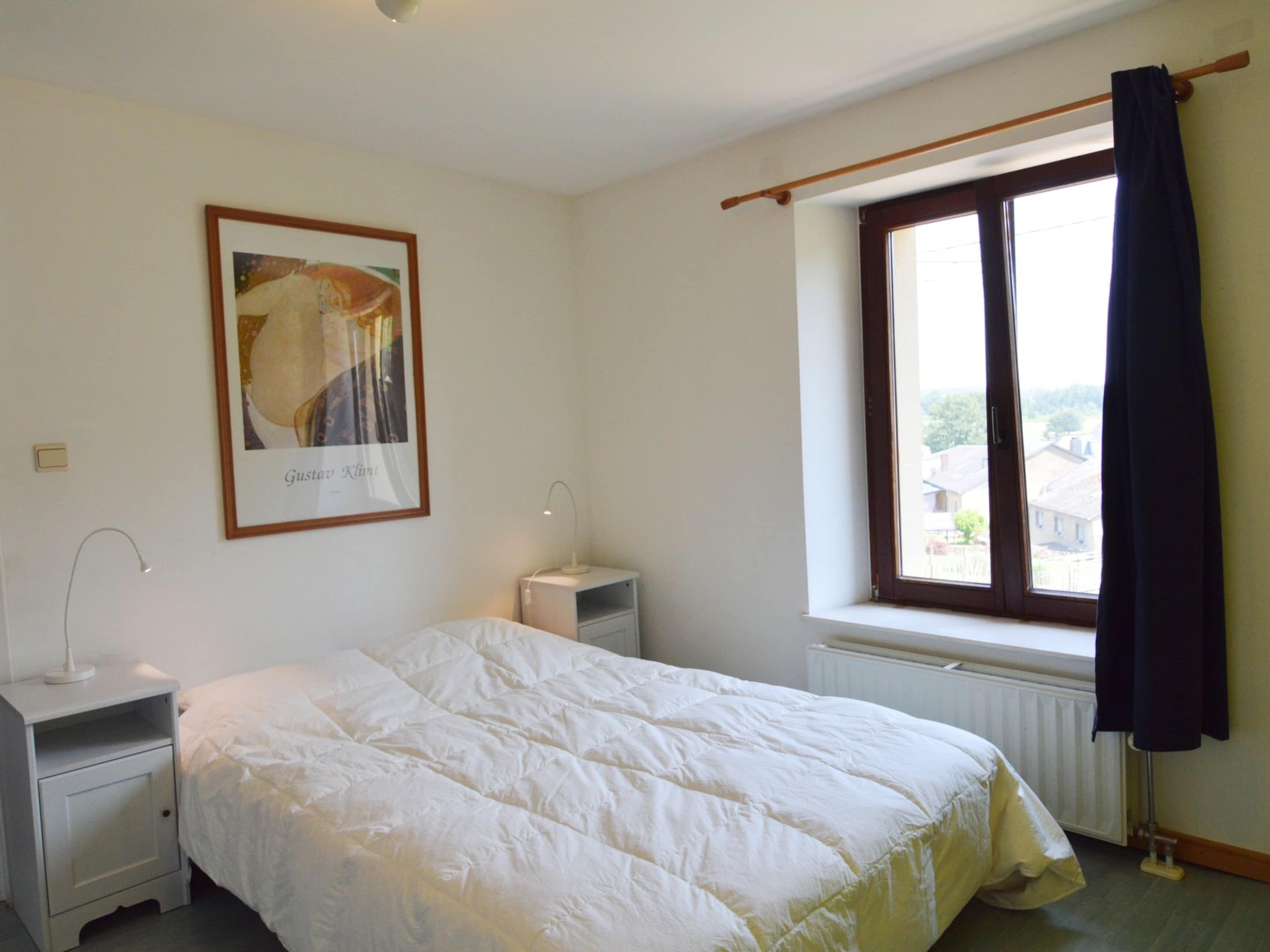Een slaapkamer met een bed, nachtkastje, lamp, raam met gordijnen en een lijst met foto aan de muur.