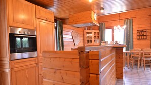A wooden cabin interior with a kitchen area featuring a Bosch oven, wooden cabinets, a heart-shaped wall decoration, and a dining area with chairs and a table. - Scenic Chalet in Ovifat (Waimes)