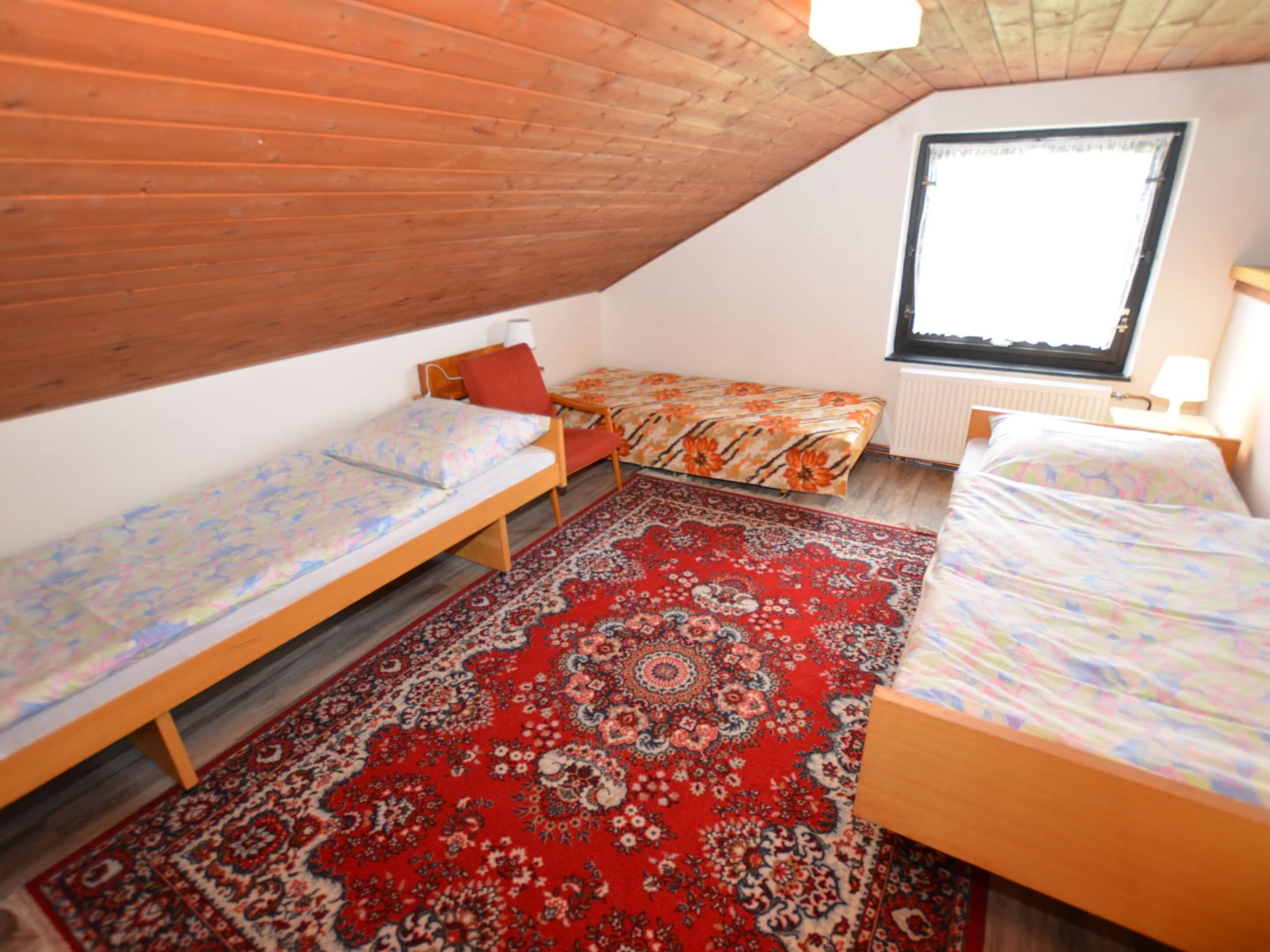 A small, cozy attic bedroom with three single beds, a large red patterned rug, and a window with a view of the sky.