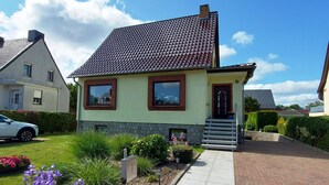 Une maison à deux étages, avec un toit en tuiles, un porche à l’avant avec des marches et un petit jardin agrémenté de fleurs et d’arbustes.