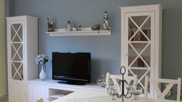 Une pièce avec un téléviseur posé sur un meuble blanc, une petite table sur laquelle se trouve un vase de fleurs, et des meubles blancs disposés contre un mur gris.