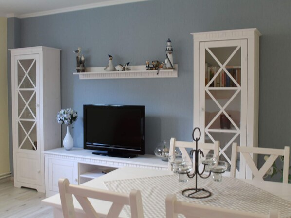 Una habitación con televisión sobre un mueble blanco, una mesita con un jarrón de flores y muebles blancos frente a una pared gris.