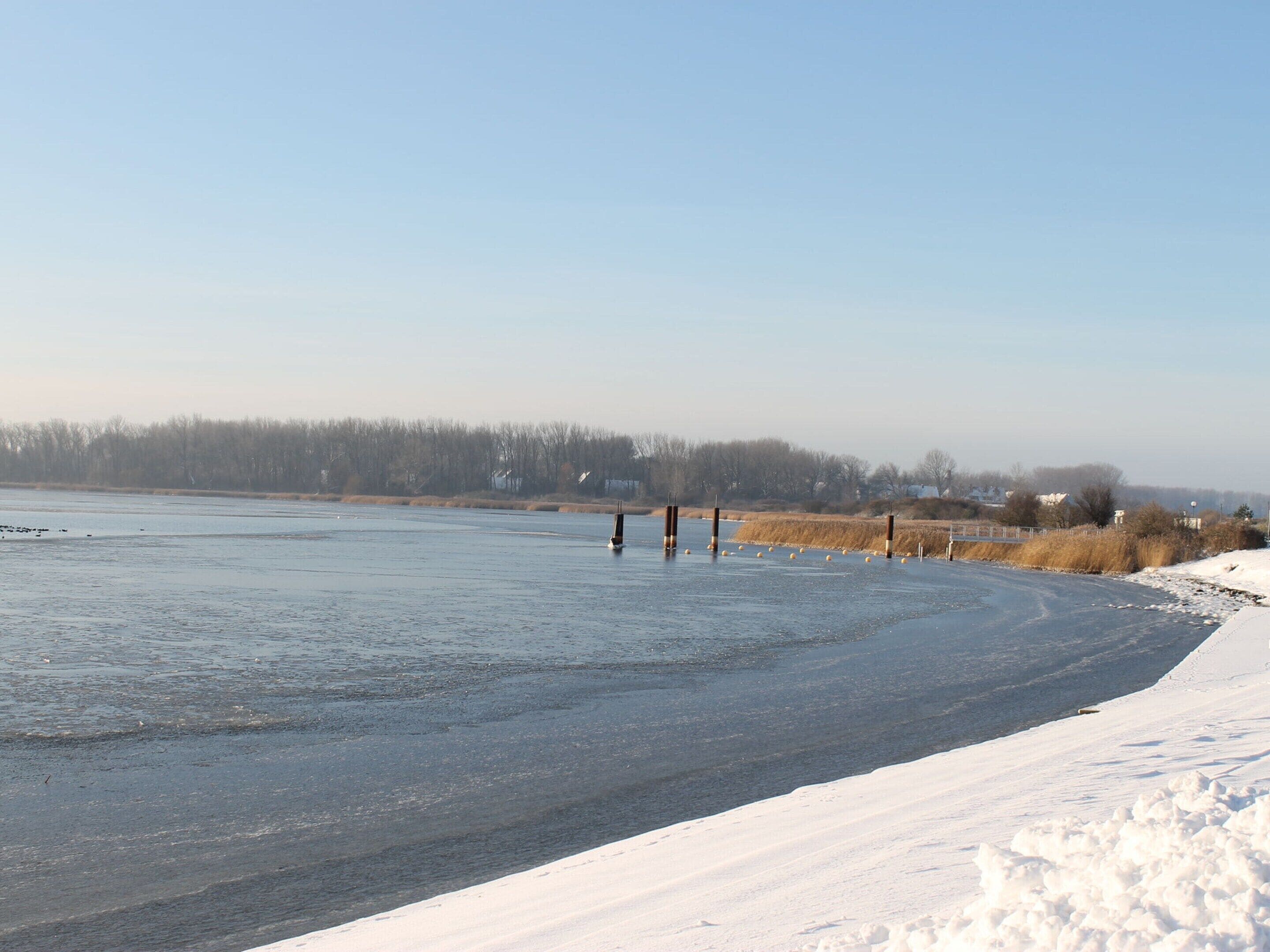 A frozen river with a sailboat, snow-covered banks, and a distant tree line.