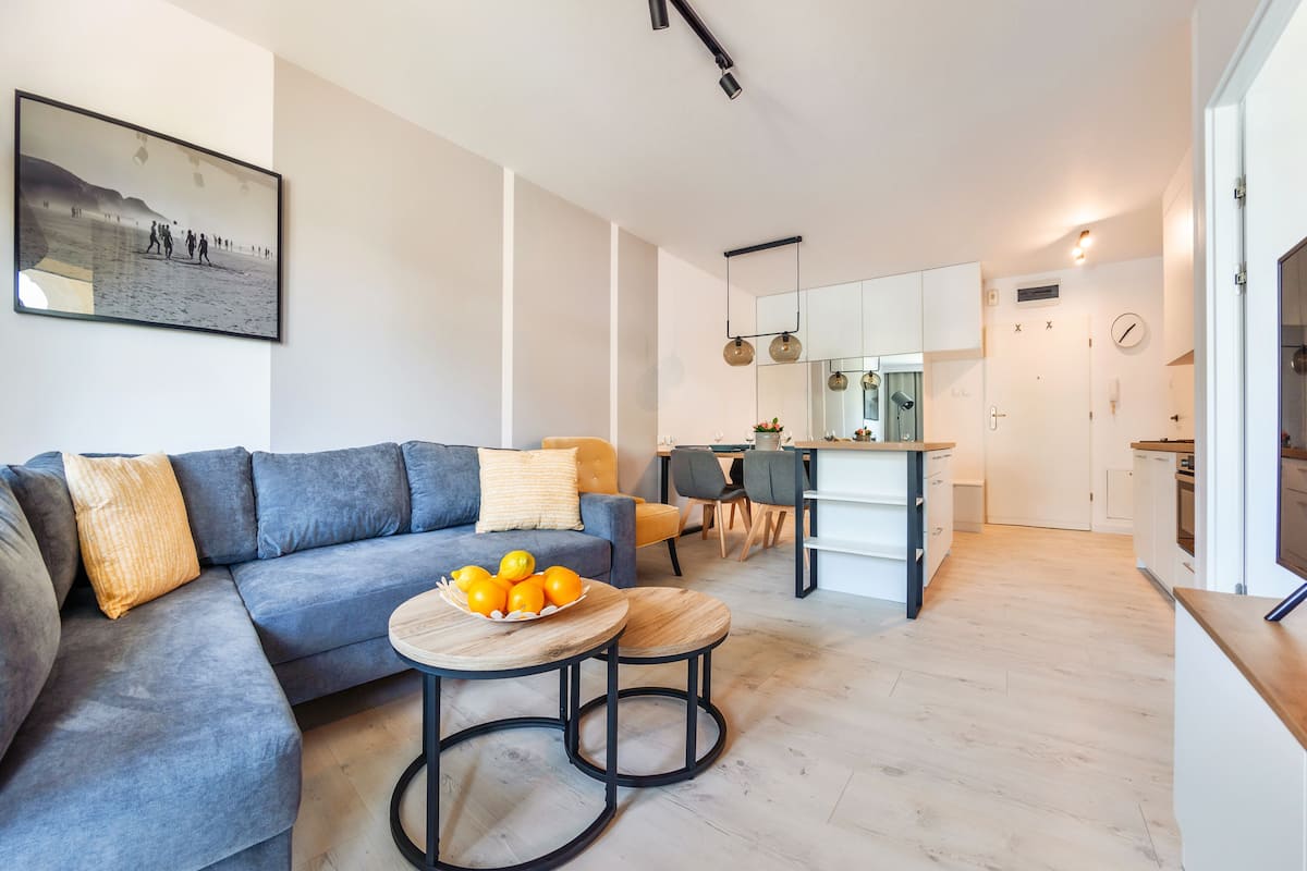 A modern living room with a grey sofa, a wooden coffee table with oranges, and a dining area with a kitchen in the background.
