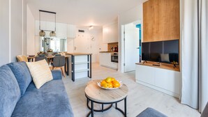 A modern living room with a sofa, a coffee table with fruit, a TV, and a kitchen area with cabinets and a dining table. - Apartamenty Sun & Snow Zielone Tarasy (Kolobrzeg)