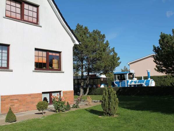 Une maison à deux étages, avec une base en briques et des murs supérieurs blancs, un petit jardin agrémenté de divers arbustes, et un train bleu et blanc en arrière-plan.
