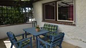 An outdoor patio with a table and chairs, a window with curtains, and a view of greenery. - Peaceful Bungalow in Diedrichshagen near Baltic Sea (Kroepelin)