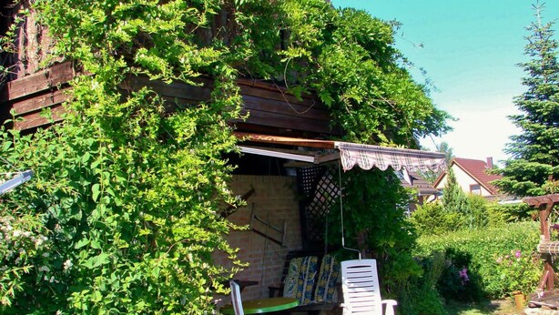 A wooden house with a covered porch, a swing, and a garden area with chairs. - Wohnung an der Ostseekuste (Wendorf)
