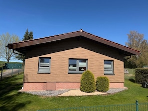 Une maison de plain-pied avec un toit brun, deux fenêtres et un petit jardin devant, agrémenté de deux arbustes.