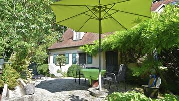 A patio area with a table and chairs under a large umbrella, surrounded by greenery and a house in the background.