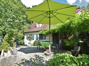 A patio area with a table and chairs under a large umbrella, surrounded by greenery and a house in the background.