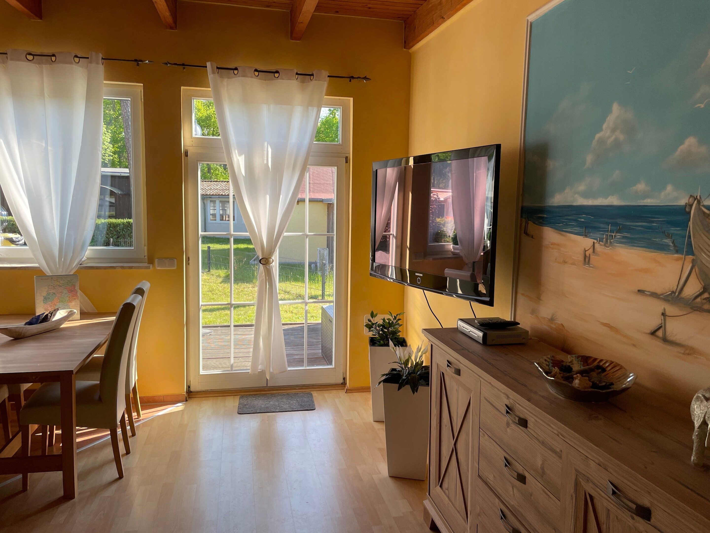 A room with a wooden dining table, chairs, a TV mounted on the wall, and a painting of a beach scene.