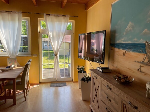 A room with a wooden dining table, chairs, a TV mounted on the wall, and a painting of a beach scene.