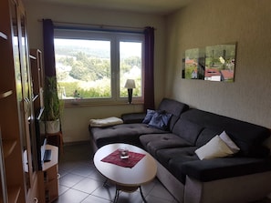A living room with a sofa, a small table, a lamp, and a window with a view of greenery and buildings.