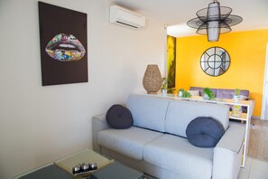 Un salon moderne avec un canapé bleu clair, deux coussins noirs, une table basse en verre, un vase décoratif et un climatiseur fixé au mur.