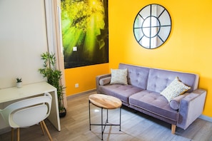 Une pièce avec un canapé violet, un bureau blanc, un petit tabouret en bois et un grand tableau abstrait accroché au mur.