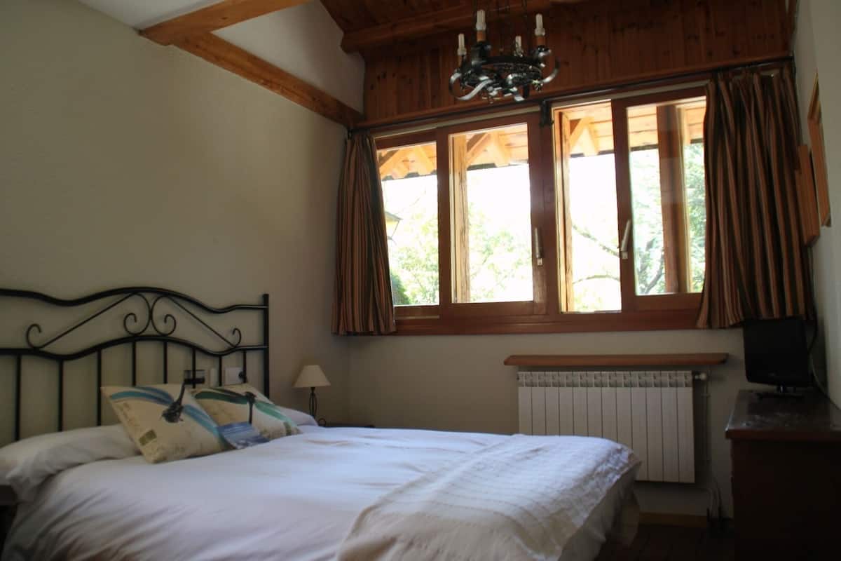 Un dormitorio con cama, ventana con cortinas, radiador y televisor sobre un soporte de madera.