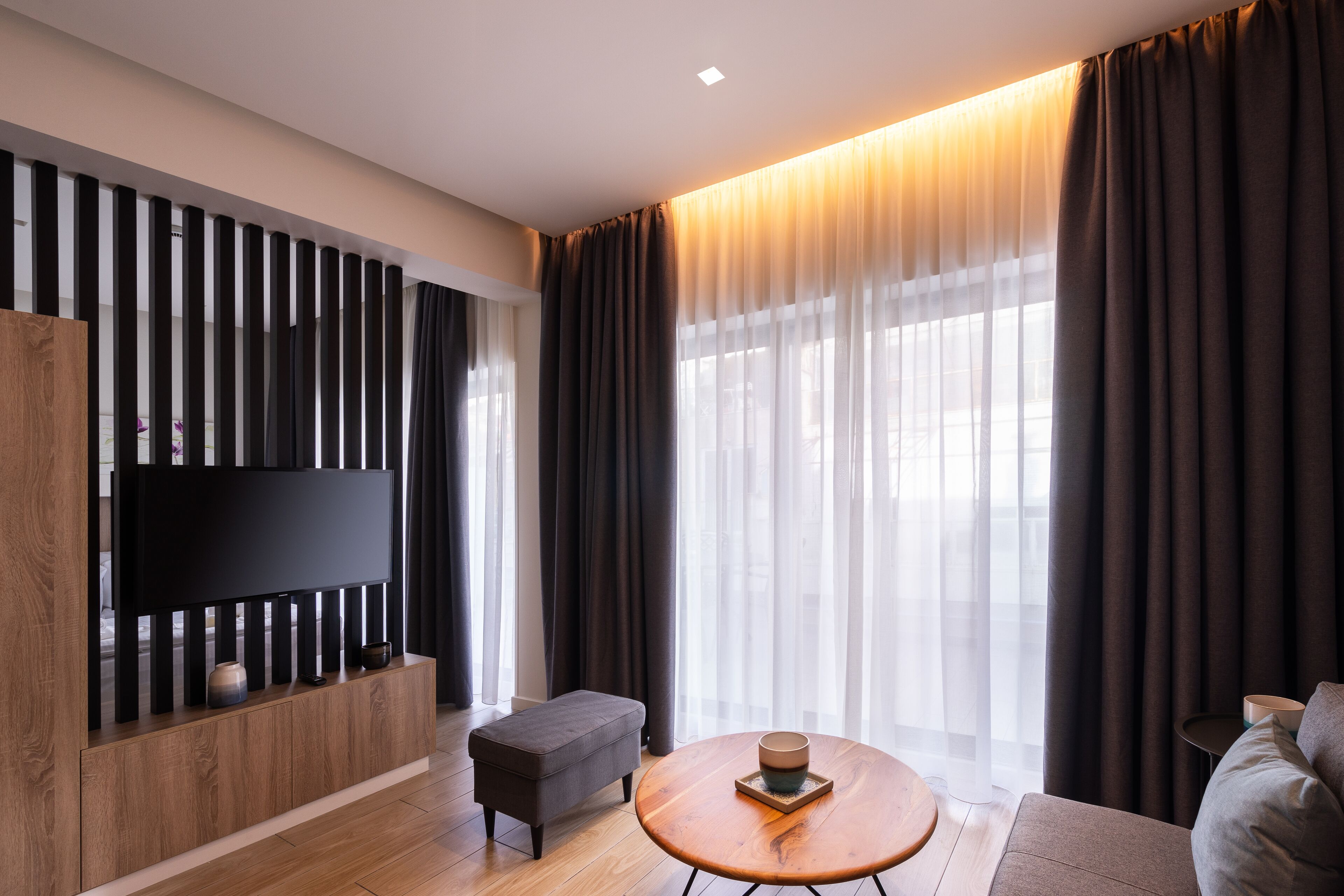 A modern living room with a flat-screen TV mounted on a wooden panel, a round wooden coffee table, a grey sofa, and a large window with sheer curtains.