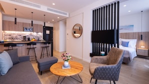 Una sala de estar moderna con sofá, sillón y mesa de centro. Se observa una zona de cocina con microondas y taburetes de bar. Un televisor grande está montado en la pared, y en el fondo se ve una cama con cabecero.