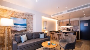 Una sala de estar moderna con un sofá, una mesa de centro y una silla. Hay una zona de cocina con microondas y taburetes de bar.