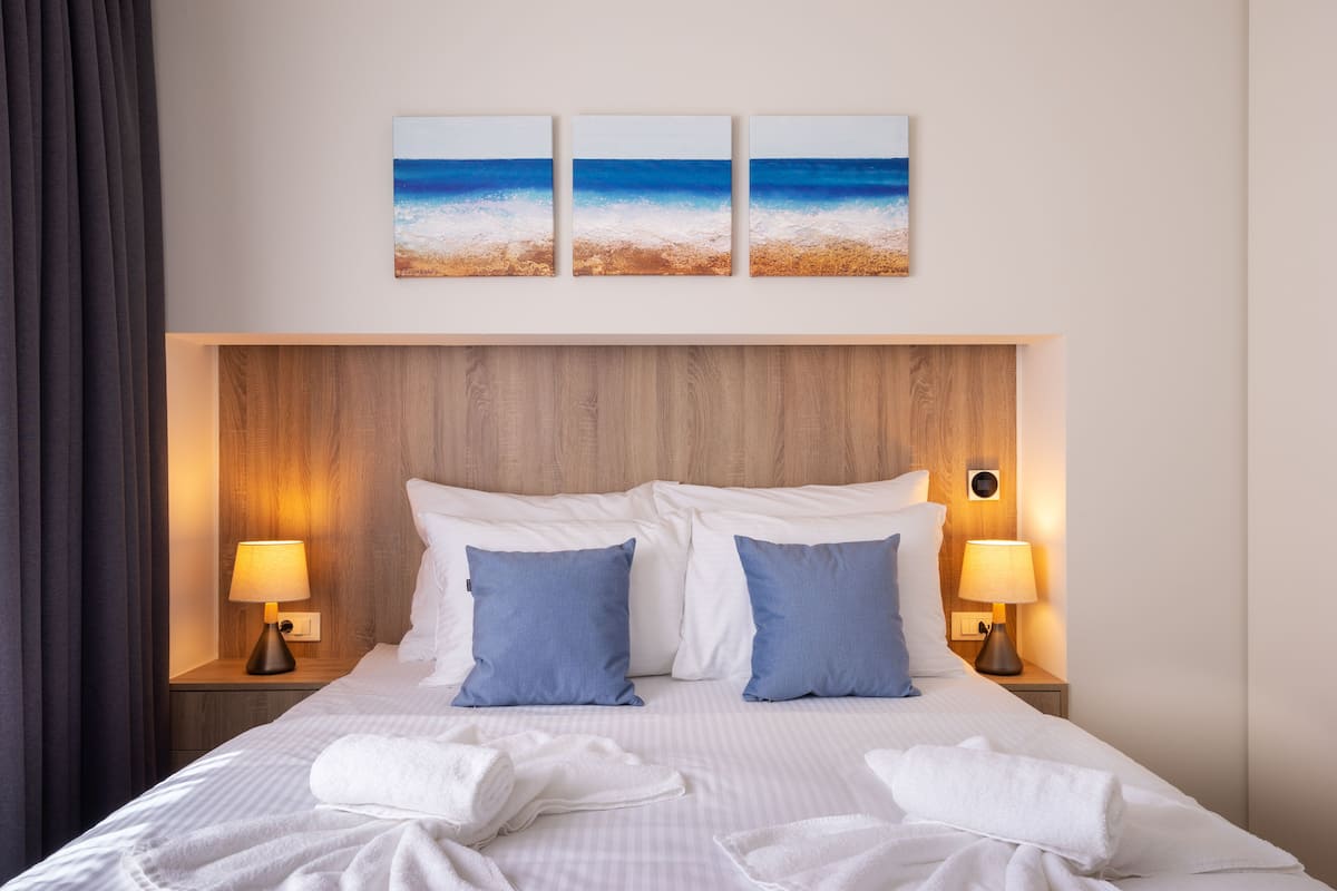 Een hotelkamer met een houten hoofdeinde, twee bedlampjes, een bed met witte lakens, twee blauwe kussens en drie strandafbeeldingen aan de muur.