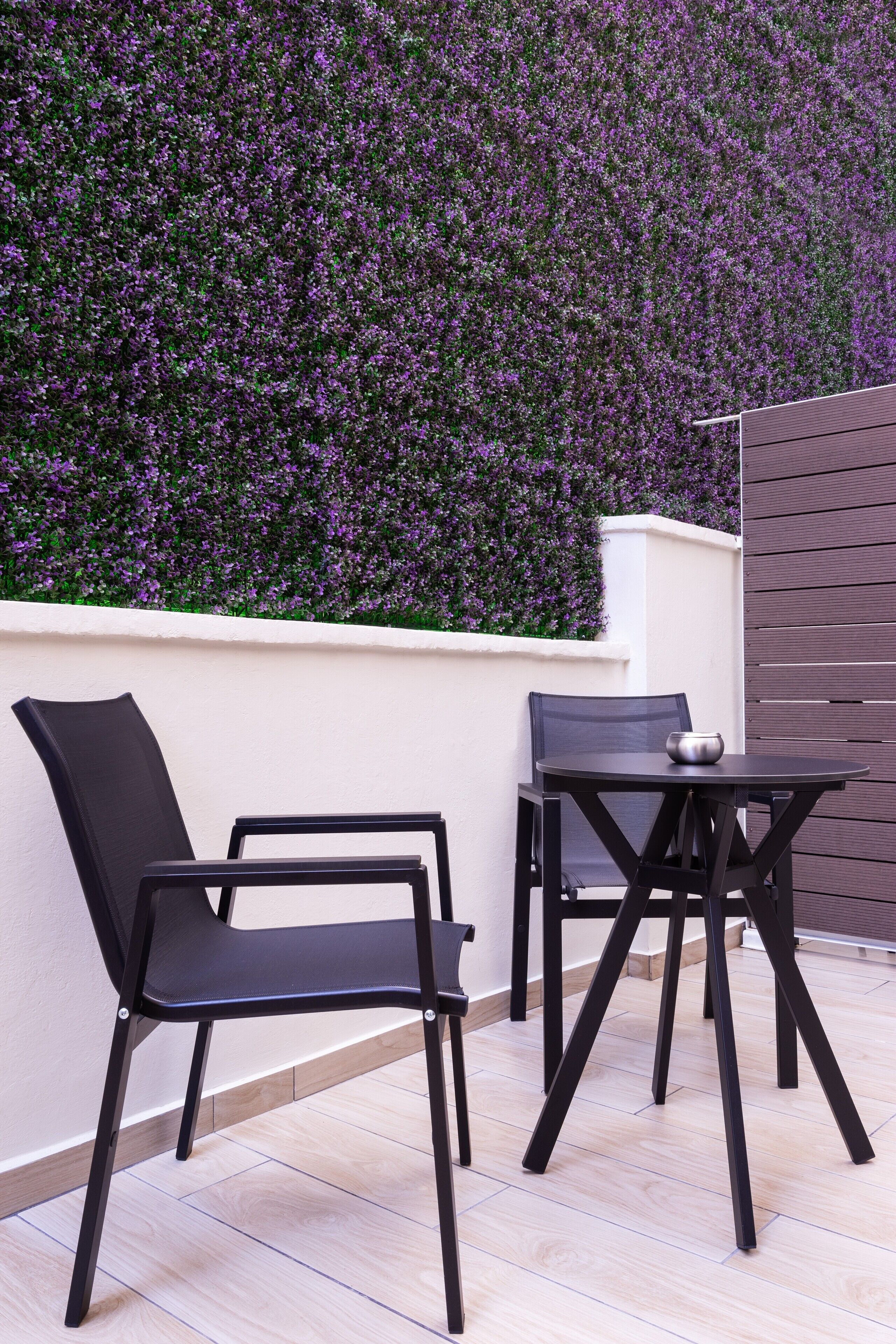 Un patio con due sedie nere in metallo e un tavolo, una parete bianca e una parete ricoperta di fiori viola.