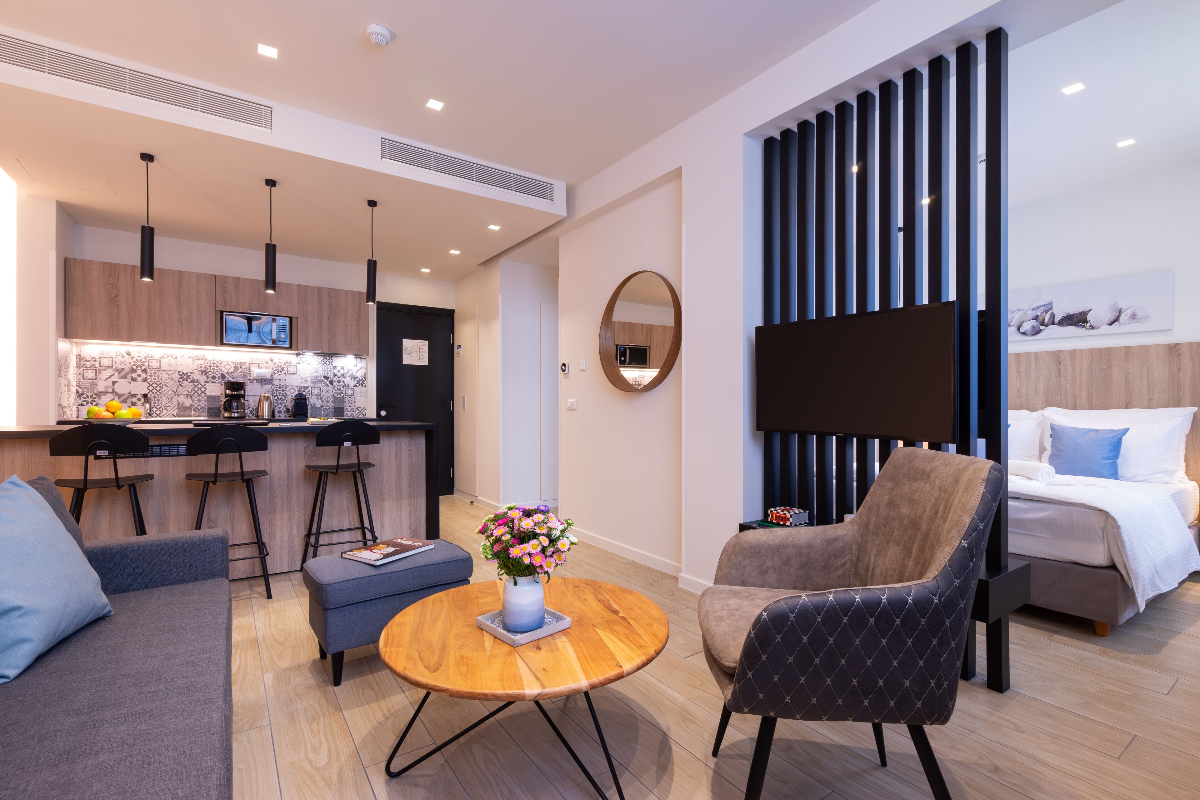 Een moderne woonkamer met een bank, fauteuil en salontafel. Er is een keukenhoek met een televisie en barstoelen. Een grote spiegel hangt aan de muur, en er is een bed in de achtergrond te zien.