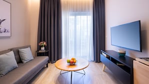 Una sala de estar moderna con un sofá, una mesa redonda con fruta, un televisor de pantalla plana y un amplio ventanal con cortinas.