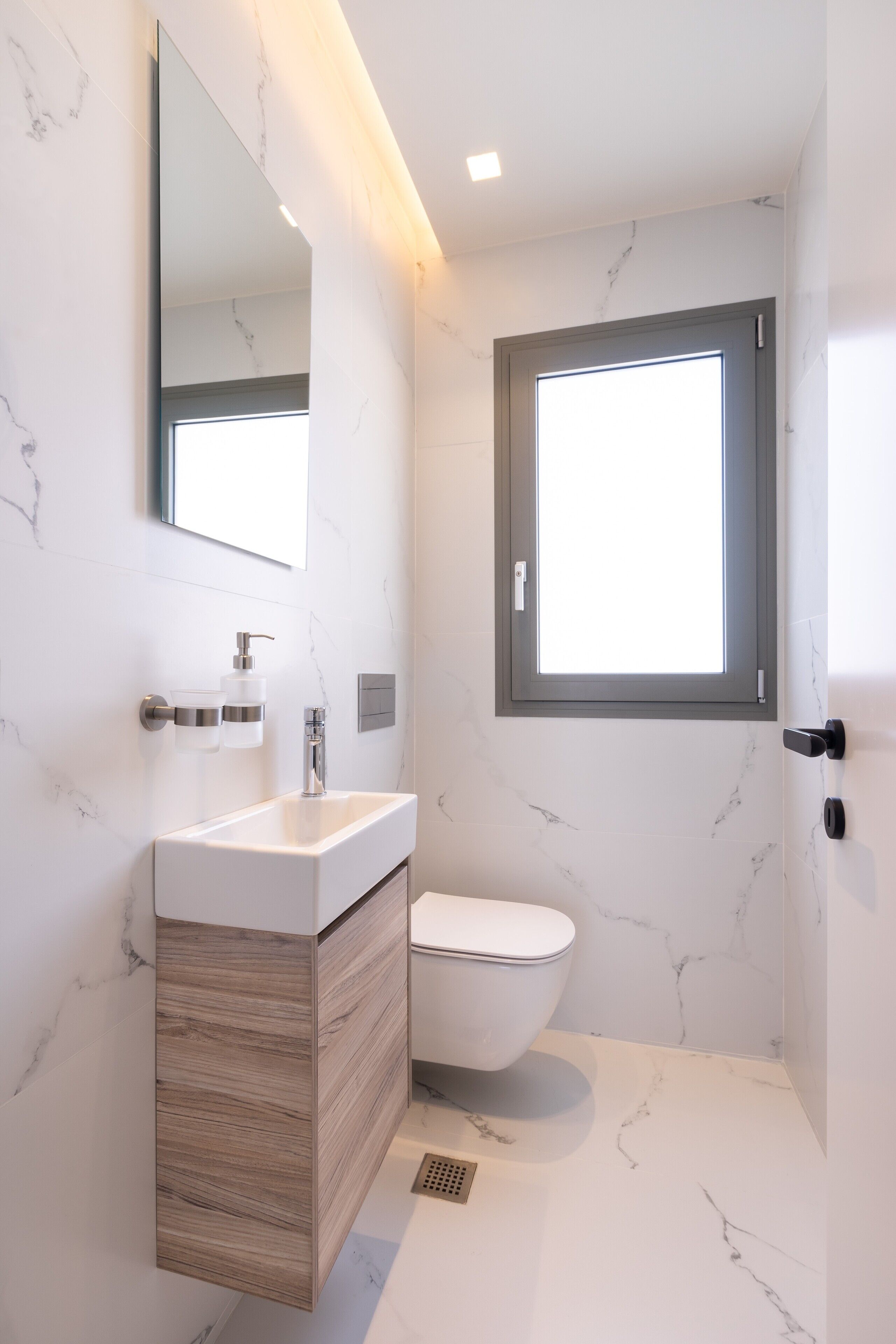 A modern bathroom with a marble wall, a wooden vanity, a white sink, a toilet, and a window.