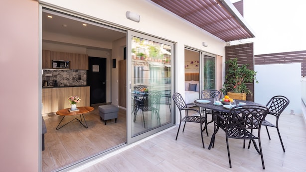 Un patio moderno con una mesa y sillas de comedor, un pequeño rincón de estar con un sofá, y una cocina visible a través de una puerta de vidrio.
