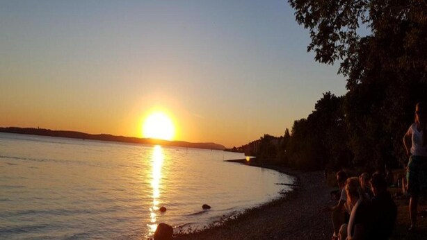A sunset over a calm body of water with a beach and trees in the background. - Spacious Apartment near Lake Constance with Covered Balcony (Bermatingen)