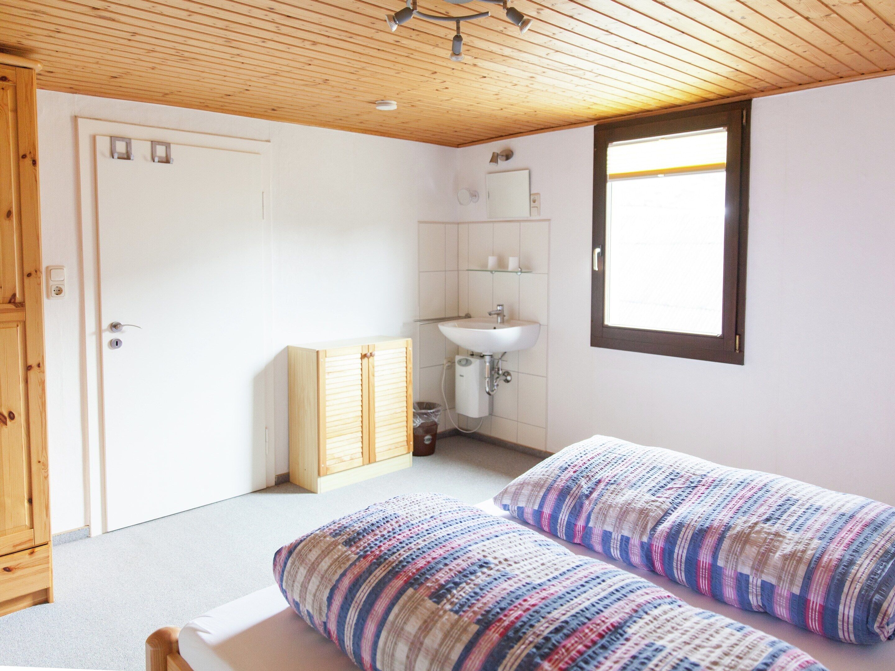 A bedroom with two beds, a wooden wardrobe, a bathroom with a sink and mirror, and a window providing natural light.