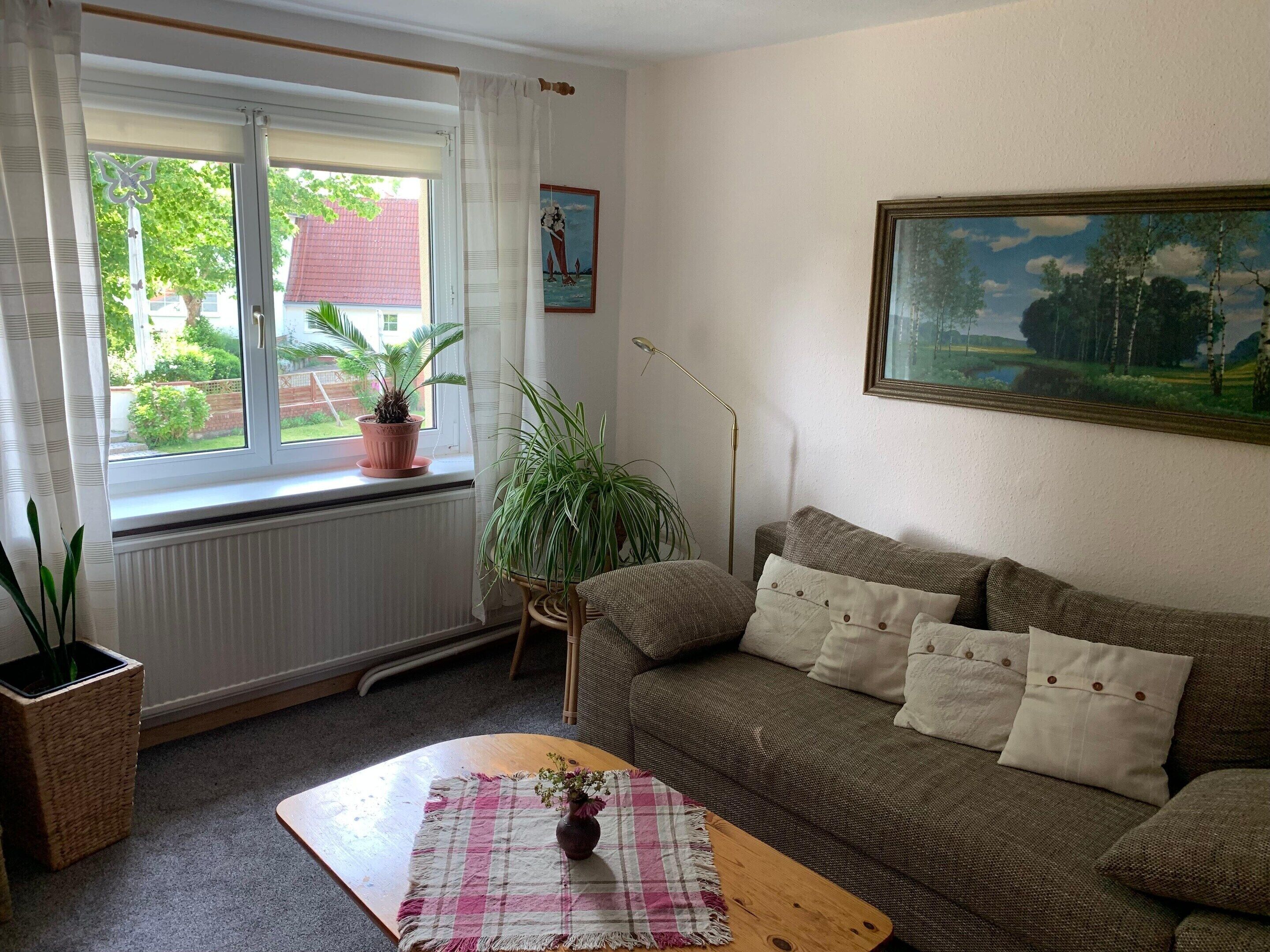 Un salon chaleureux avec un canapé, une table basse surmontée d’un vase, une fenêtre avec des rideaux et un tableau accroché au mur.