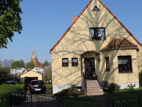 Egy kétszintes ház, piros tetővel, egy kis verandával, és a homlokzatán jól láthatóan szerepel a „21”-es házszám.
