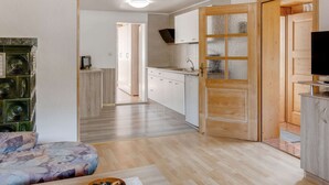 Ein moderner, gut beleuchteter Raum mit Holzbalkendecke, einer Couch, einem Fernseher und einer Küchenzeile mit weißen Schränken und einer Holztür.