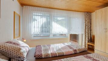 A bedroom with two beds, wooden ceiling, large windows with sheer curtains, and a wooden wardrobe.