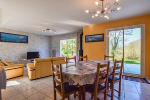 A living room with a sofa set, a dining table with chairs, a TV, and a large window with a view of greenery. - Coastal Retreat in Plurien (Plurien)