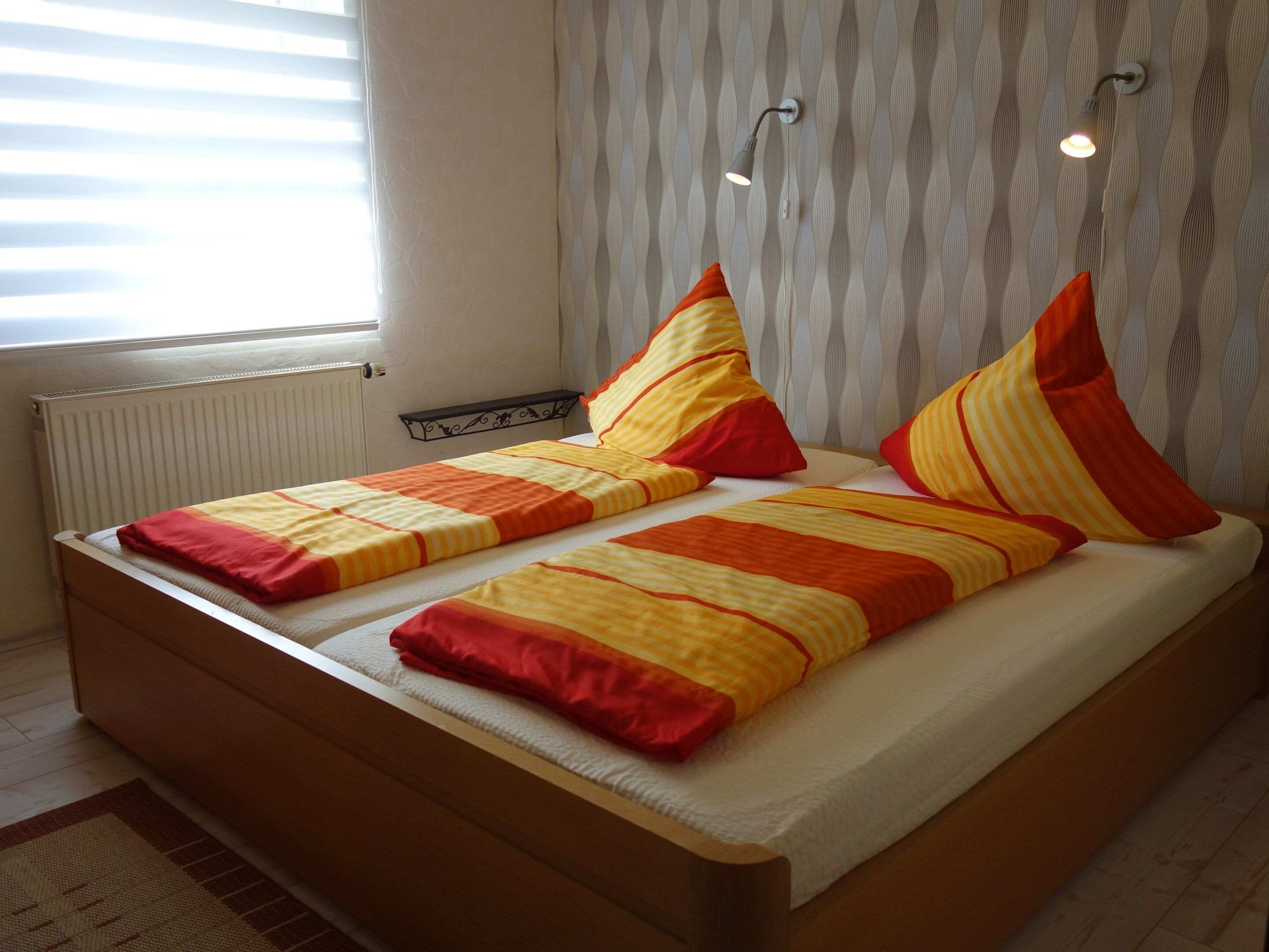 A double bed with striped bedding and red pillows, a bedside table, a radiator, and a window with blinds.