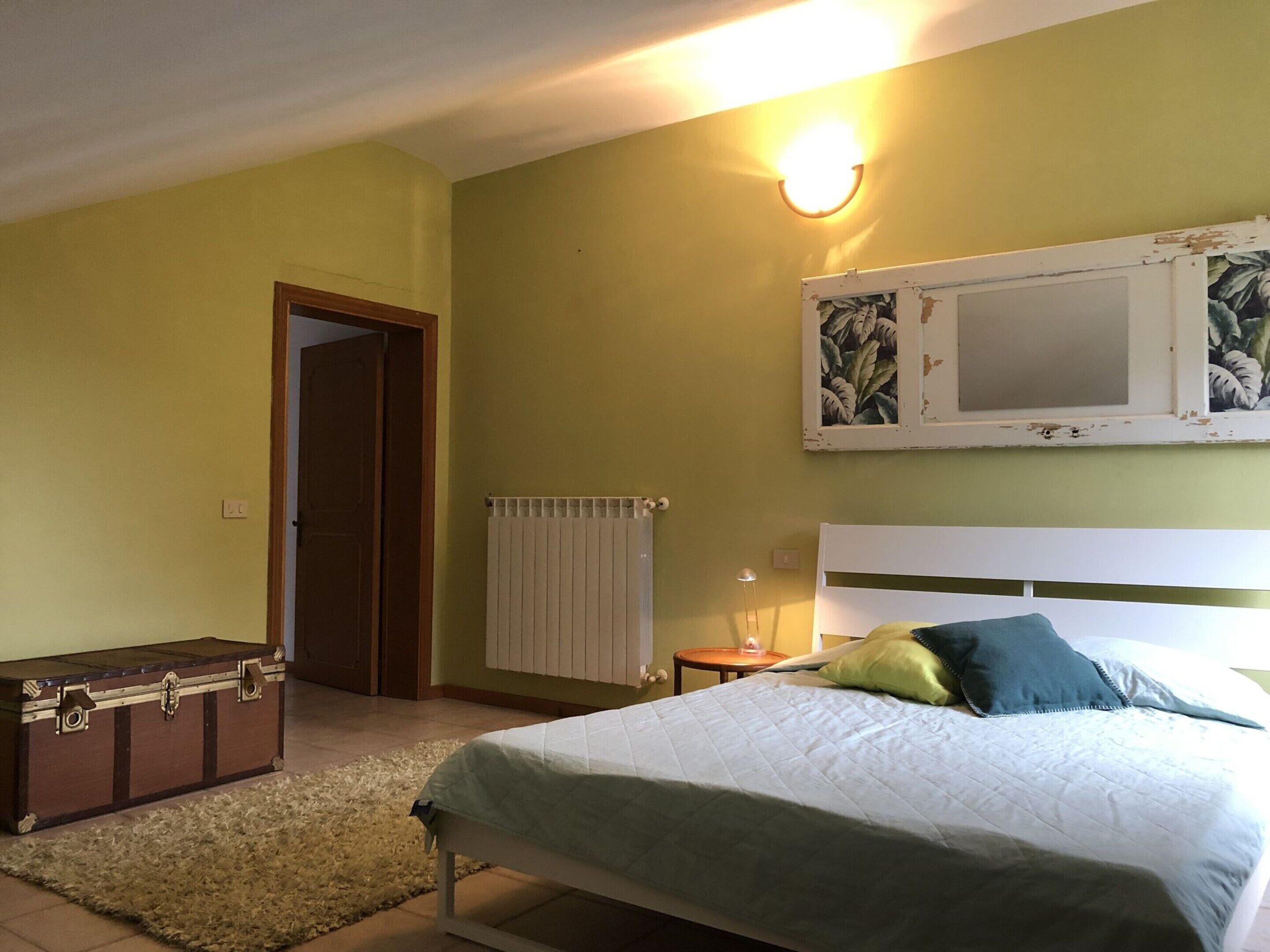 Ein Schlafzimmer mit Bett, Nachttisch, Lampe, Heizkörper, Tür und Koffer.